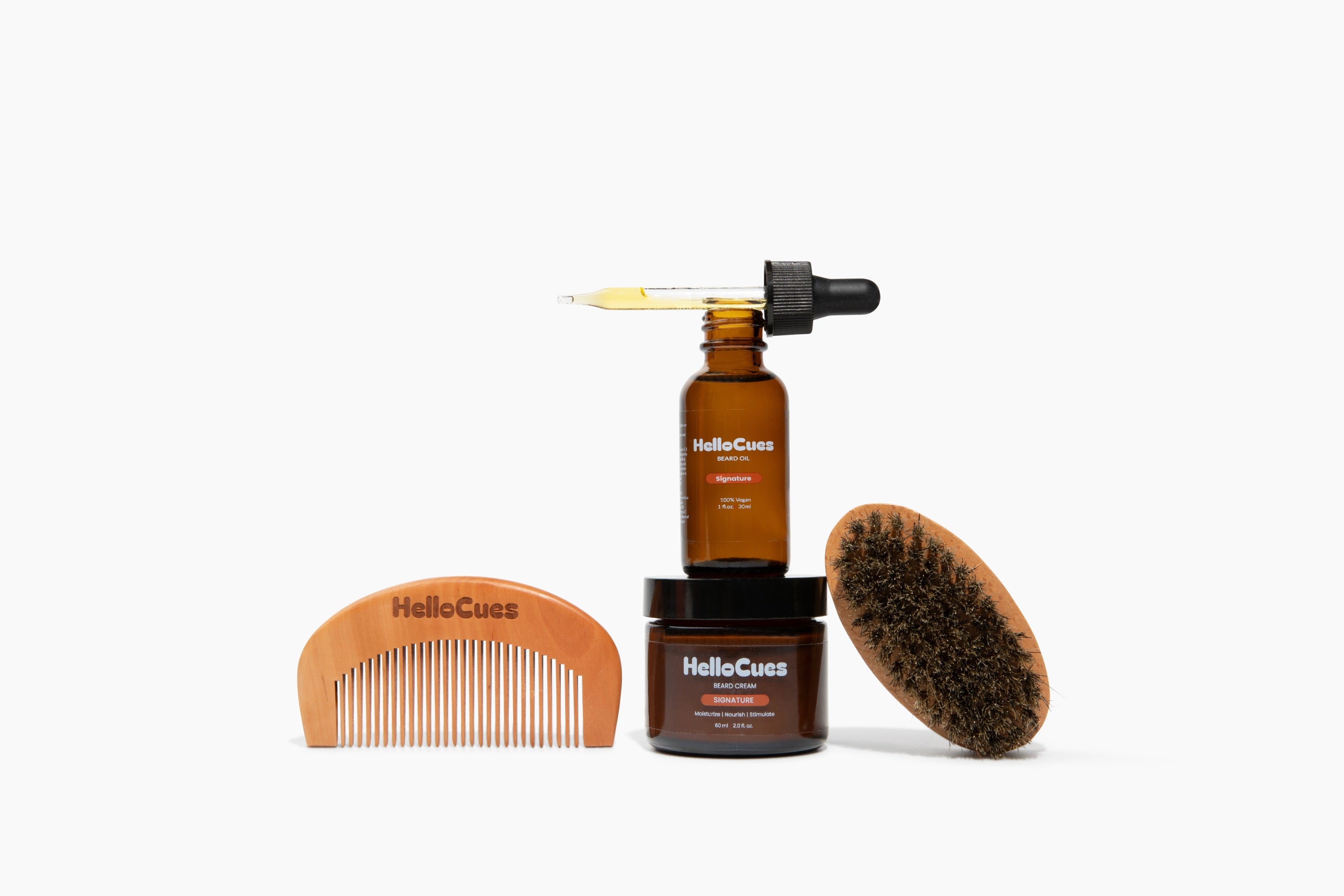 HelloCues Beard Care Deluxe: Signature beard oil in an amber dropper bottle, a 2 oz jar of Signature beard cream, a wooden HelloCues beard comb and a boar-bristle beard brush, arranged on a plain white background.